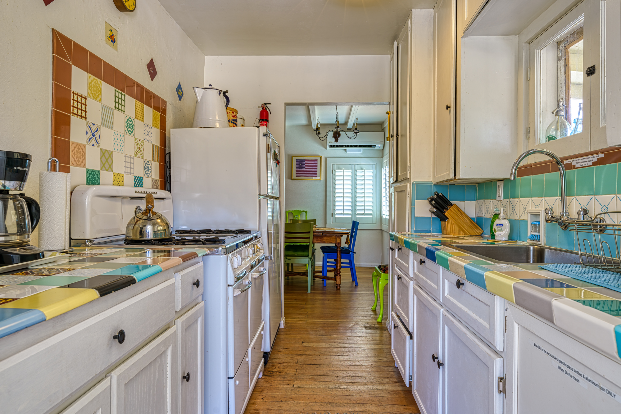 A classic old timey kitchen with tile countertops, 50s to 60s style stove and fridge with fire extinguisher on top of the fridge. Coffee pot, tea kettle, paper towel holder to the left by the stove and fridge with kitchen sink, knives, dish rack, and trash recycling combo to the right.