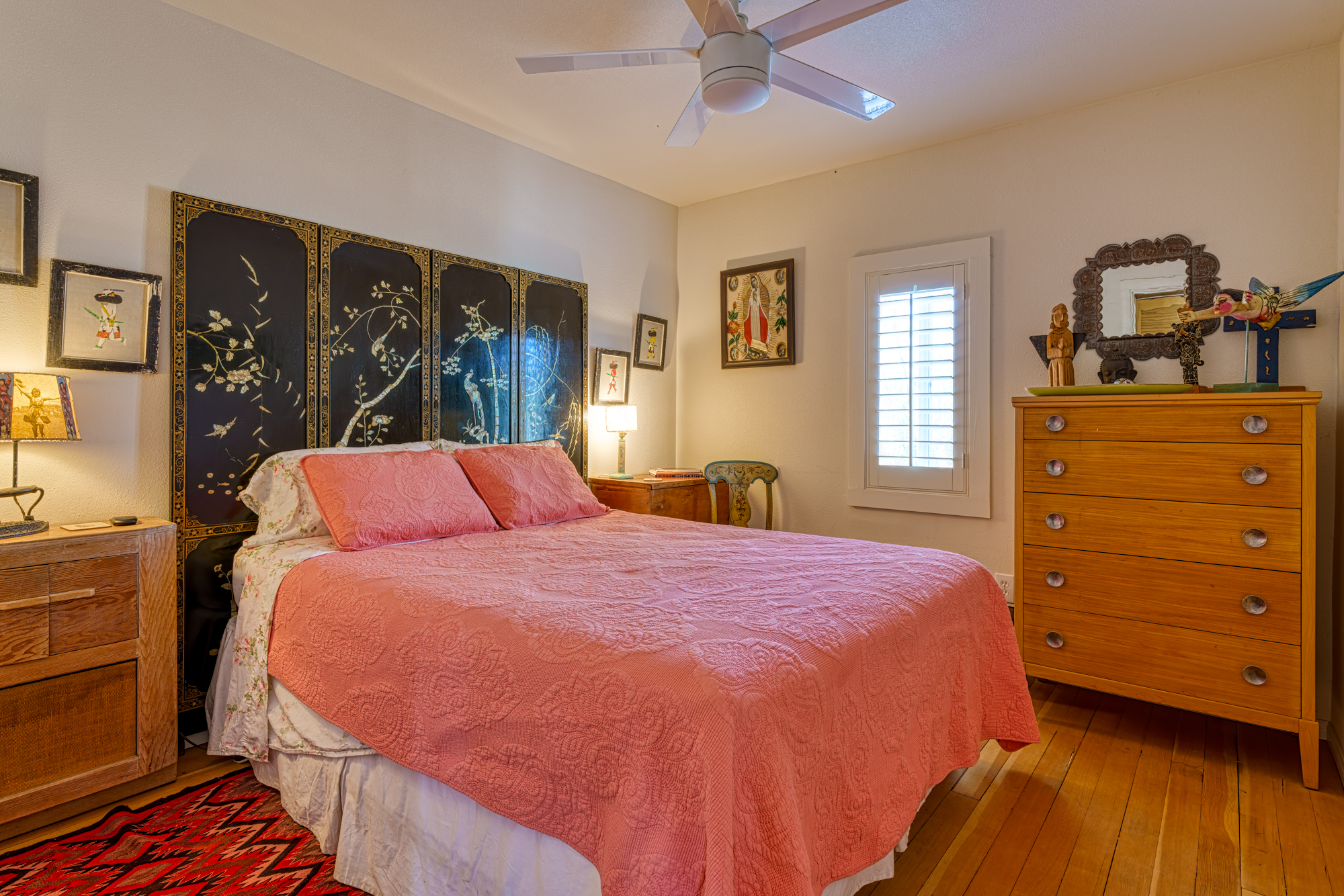 Main bedroom containing a large bed with medium sized wardrobe that has various memorabilia on top of it, two nightstands, a single chair to the right of the bed, various painting and decor hanged from the walls, a rug, and a ceiling fan in the center of the ceiling.