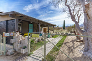 View of Agave Art House's front porch with eclectic decor, including a 'Killer Dogs' sign, set against a backdrop of desert flora and expansive skies.