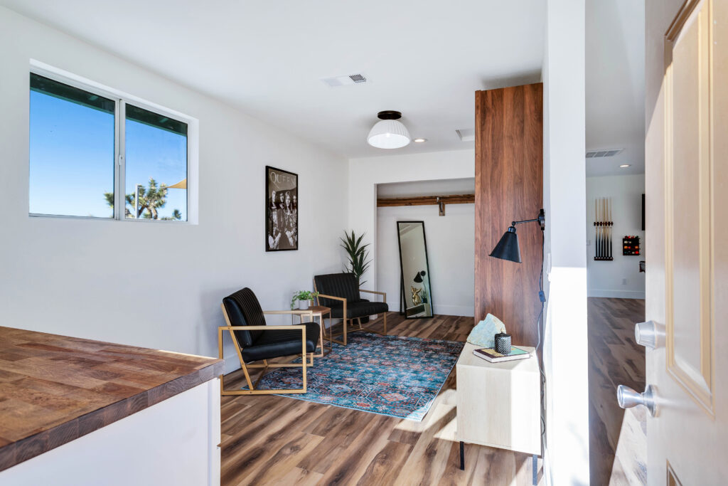 a Sleeping Area with a Full-sized Murphy Bed, Modern Chairs, a Wooden Side Table, and a Colorful Rug. the Room Includes a Large Mirror, a Wall-mounted Lamp, and a Plant, with Natural Light Streaming Through the Window. the Decor is Stylish and Inviting.