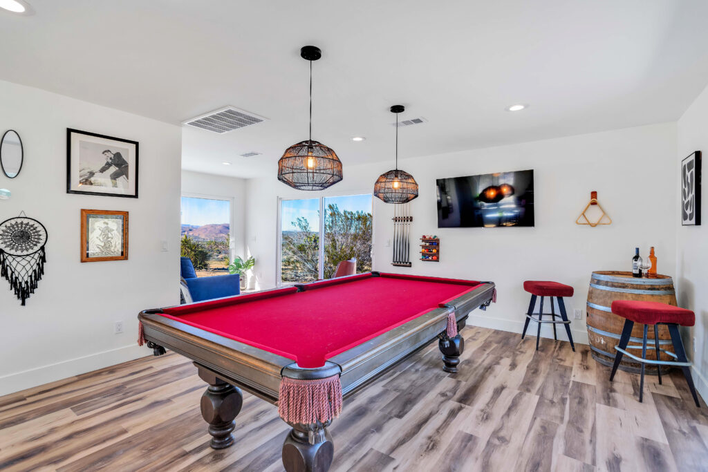 a Game Room with a Red Felt Pool Table, Stylish Pendant Lights, a Wall-mounted Tv, Bar Stools, and a Wine Barrel Table. the Room Also Features Large Windows with Desert Views and Various Decorative Elements Like Framed Pictures and a Dreamcatcher.