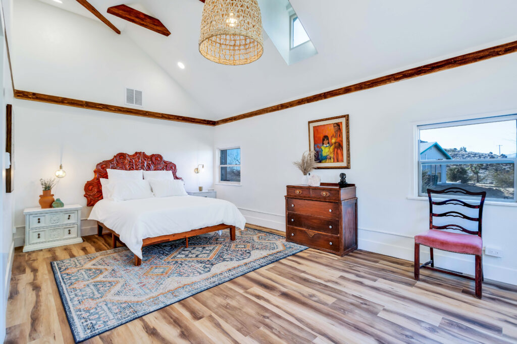 Large Open Bedroom with Cathedral Ceilings and Multiple Windows with Views to the Desert Outside. Room Features a Queen Bed with an Ornate Headed with End Tables and Wall Sconces on Both Sides. the Room Also Has a Dresser and a Chair.