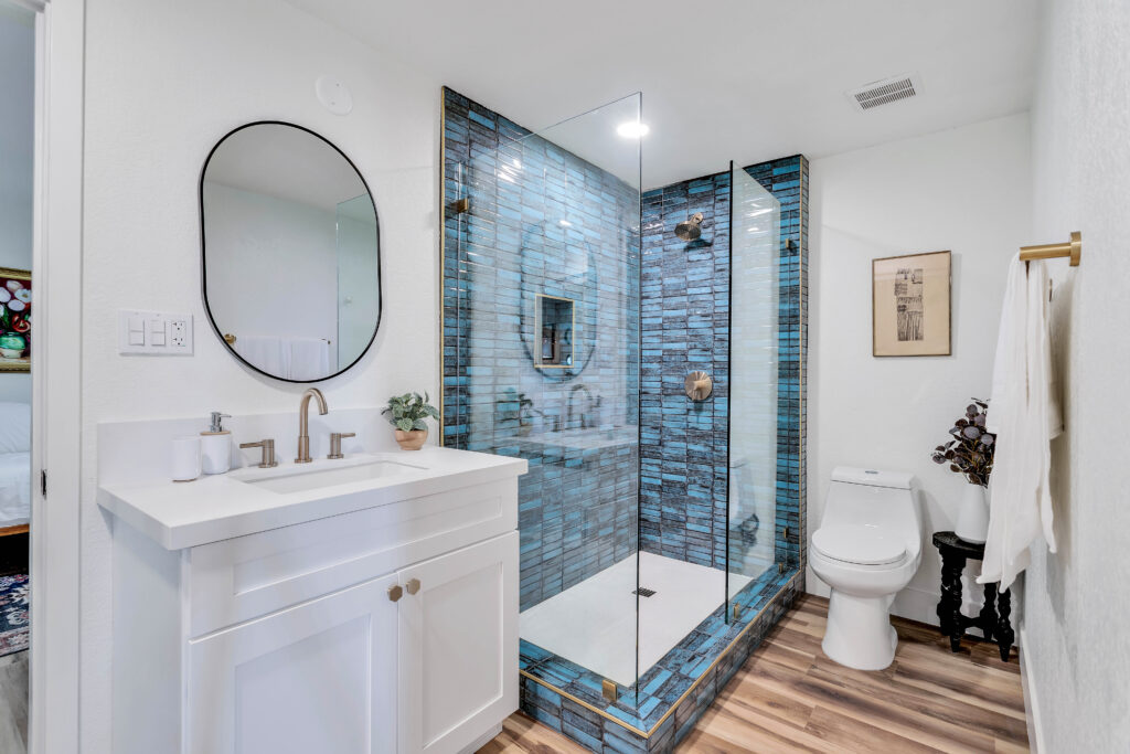 a Shared Bathroom with a White Vanity, Oval Mirror, and a Modern Glass-enclosed Shower with Blue Tiles. the Room Includes a Toilet, Towel Rack, Decorative Plant, and Artwork on the Wall. the Decor is Sleek and Inviting.