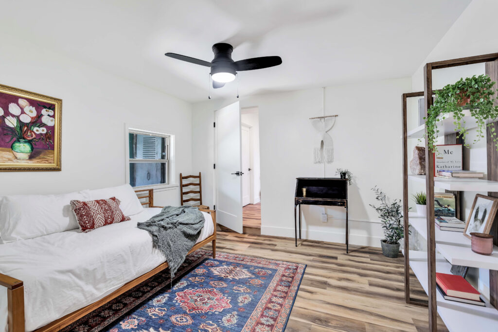 a Bedroom with a Daybed, Colorful Rug, Bookshelf, and Desk. the Room Includes Decorative Pillows, Wall Art, Plants, and a Ceiling Fan. the Space is Bright and Inviting, Ideal for Relaxation and Comfort.