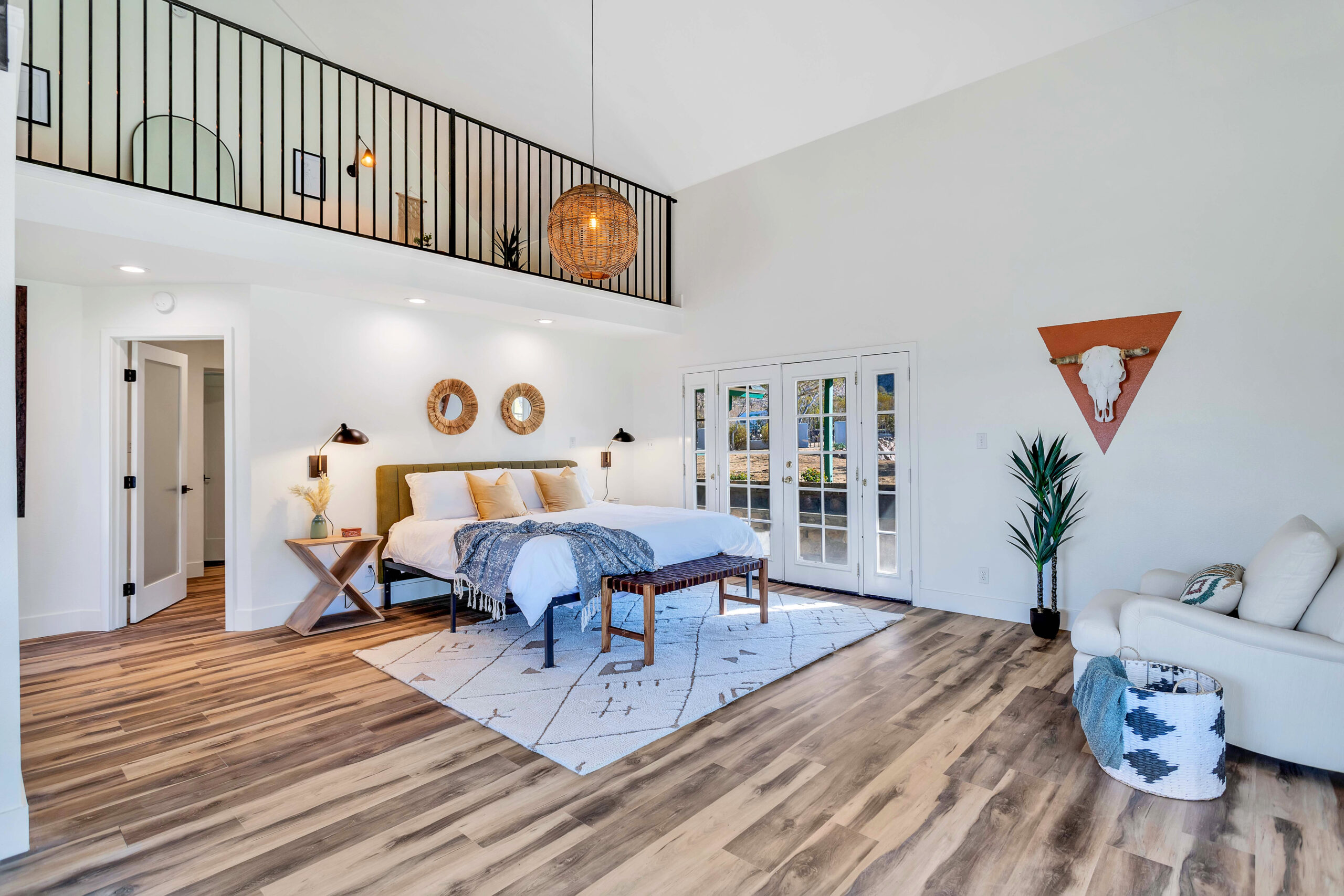 a Spacious Master Suite with a King-sized Bed, Stylish Decor, and Plenty of Natural Light. the Room Includes a White Armchair, a Bench at the Foot of the Bed, a Woven Rug, and Large French Doors. a Loft Area and Decorative Wall Elements Add to the Charm.