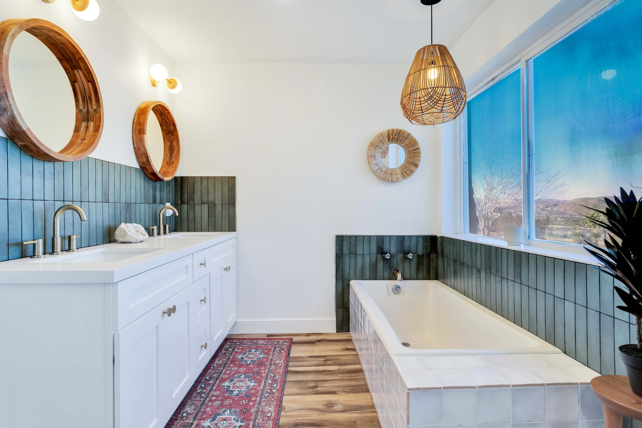 a Luxurious En-suite Bathroom with a Double Vanity, Deep Soaking Tub, and Large Window Offering Desert Views. the Space Includes Wooden-framed Mirrors, a Wicker Pendant Light, and Green Tiled Backsplash. the Decor is Elegant and Inviting.