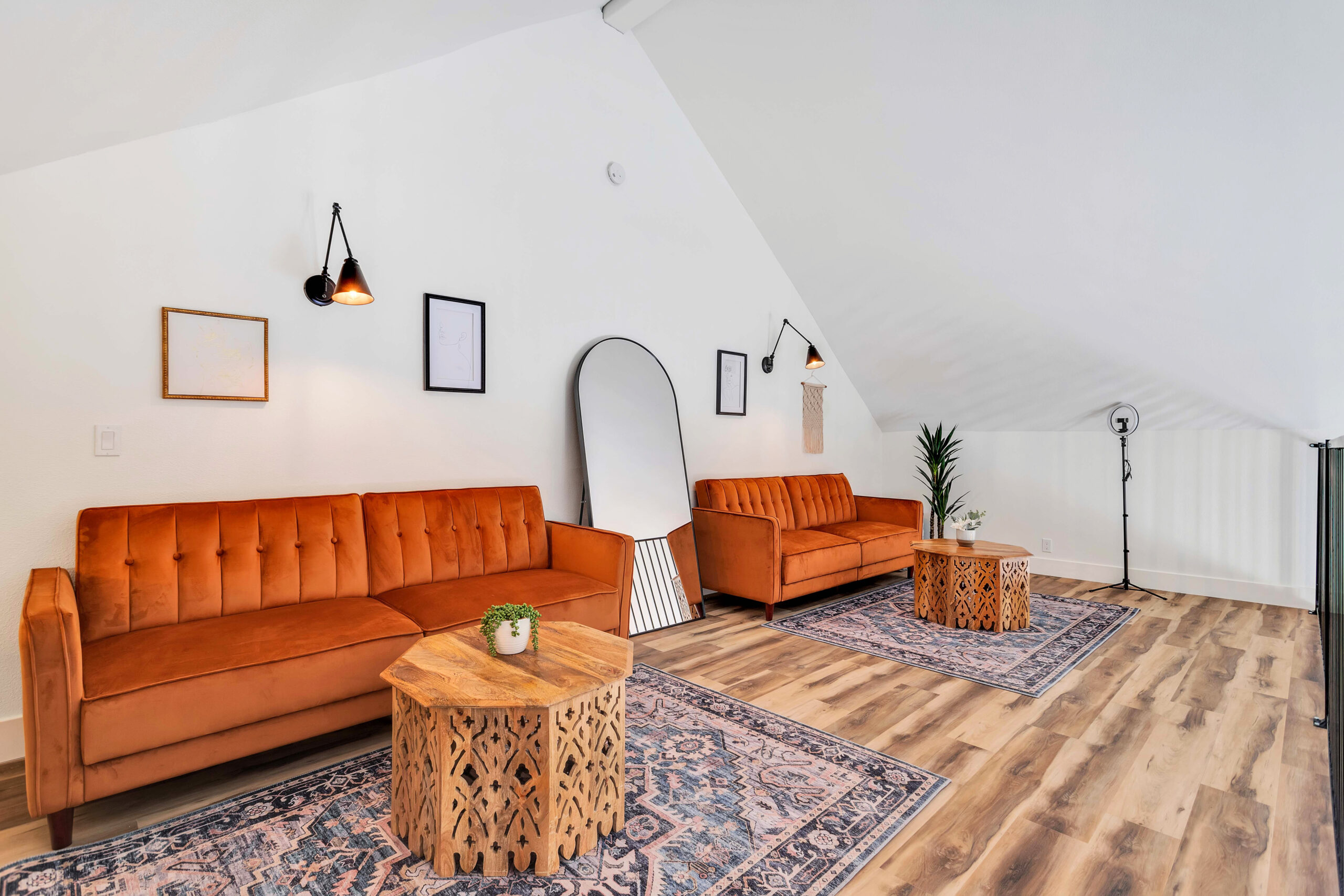 a Loft with Two Orange Sofas That Convert to Beds, Wooden Tables, Decorative Rugs, Wall Art, and a Large Mirror. the Space is Bright with a Sloped Ceiling and Stylish Decor, Providing a Cozy and Functional Area for Guests.