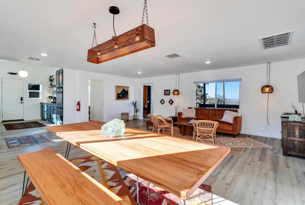 a Dining Area with a Large Wooden Table and Benches, Stylish Pendant Lighting, and Natural Light from Large Windows. the Space Includes a Cozy Seating Area with a Sofa and Wicker Chairs, a Rug, and Various Decorative Items. the Room is Bright and Welcoming.