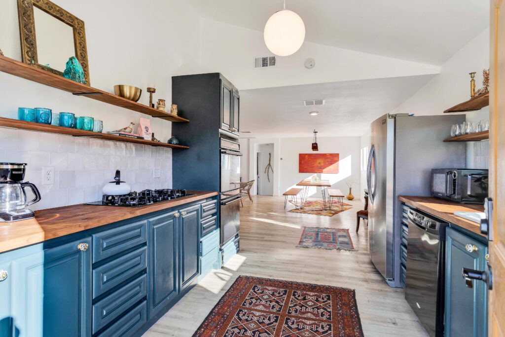 a Kitchen with Blue Cabinets, Wooden Countertops, and Open Shelving. the Space Includes a Sink Under a Window, a Gas Stove, a Dishwasher, a Refrigerator, and a Cozy Rug on the Floor. the Kitchen is Bright with Natural Light and Decorated with Plants and Decorative Items.