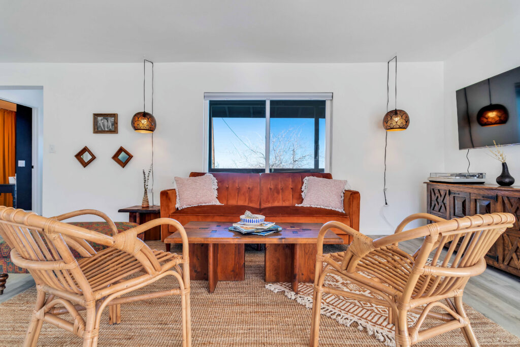 a Cozy Living Room Featuring a Burnt Orange Sofa with White Pillows, a Wooden Coffee Table, Wicker Chairs, Hanging Pendant Lights, a Wall-mounted Tv, and a Jute Rug. the Room is Decorated with Small Framed Pictures and Has a Window Showcasing a View of the Desert Landscape.