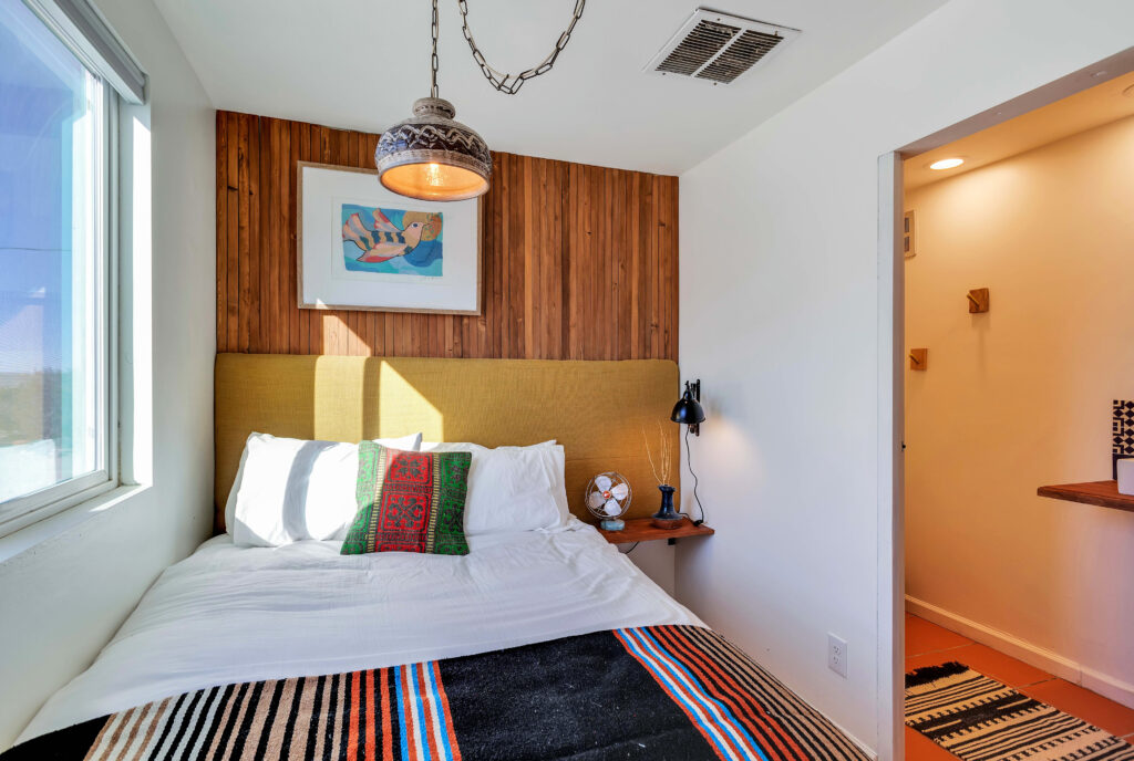 a Bedroom with a Full-sized Bed, Warm Wood Accents, a Hanging Pendant Light, and a Half Bath. the Room Includes Decorative Pillows, a Colorful Blanket, Wall Art, and a Bedside Table with a Lamp and Fan. the Space is Cozy and Inviting.