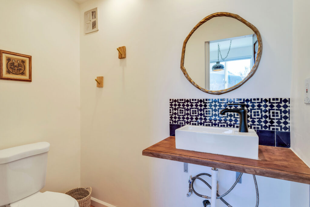 a Half Bath with a White Toilet, Vessel Sink on a Wooden Countertop, and a Round Mirror. the Decor Includes Blue Patterned Tiles, Wooden Hooks, and a Framed Artwork. the Space is Stylish and Functional.