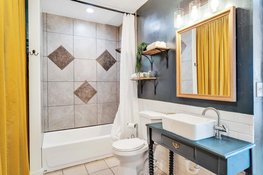 a Shared Bathroom with a Tiled Shower and Bathtub, a Stylish Vanity with a Vessel Sink, a Framed Mirror, and Open Shelves with Towels and Decor. the Room Also Serves As a Laundry Area, Combining Functionality with Charm.