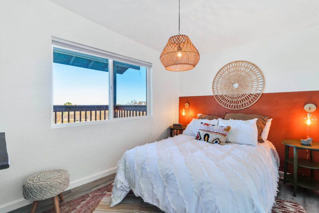 a Bedroom with a Full-sized Bed, Decorative Pillows, a Woven Wall Hanging, and Bedside Tables with Lamps. the Room Includes a Wicker Pendant Light, a Pouf, and a Window Offering Views of the Desert Landscape. the Space is Cozy and Stylish.