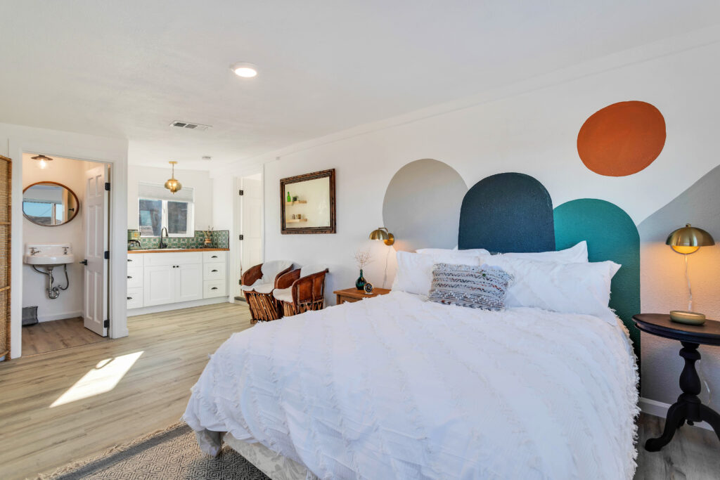 a Master Suite with a Comfortable Bed, Colorful Mural on the Wall, Cozy Seating Area, and an En-suite Bathroom. the Room Includes Decorative Pillows, Bedside Tables with Lamps, and a Modern Vanity. the Space is Bright and Inviting.