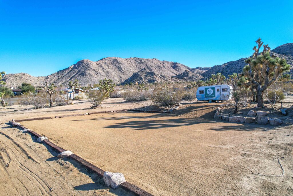 a Leveled Sandy Area for Events, with a Travel Trailer Displaying the Onaga Oasis Logo, Surrounded by Desert Landscape and Mountains Under a Clear Blue Sky.