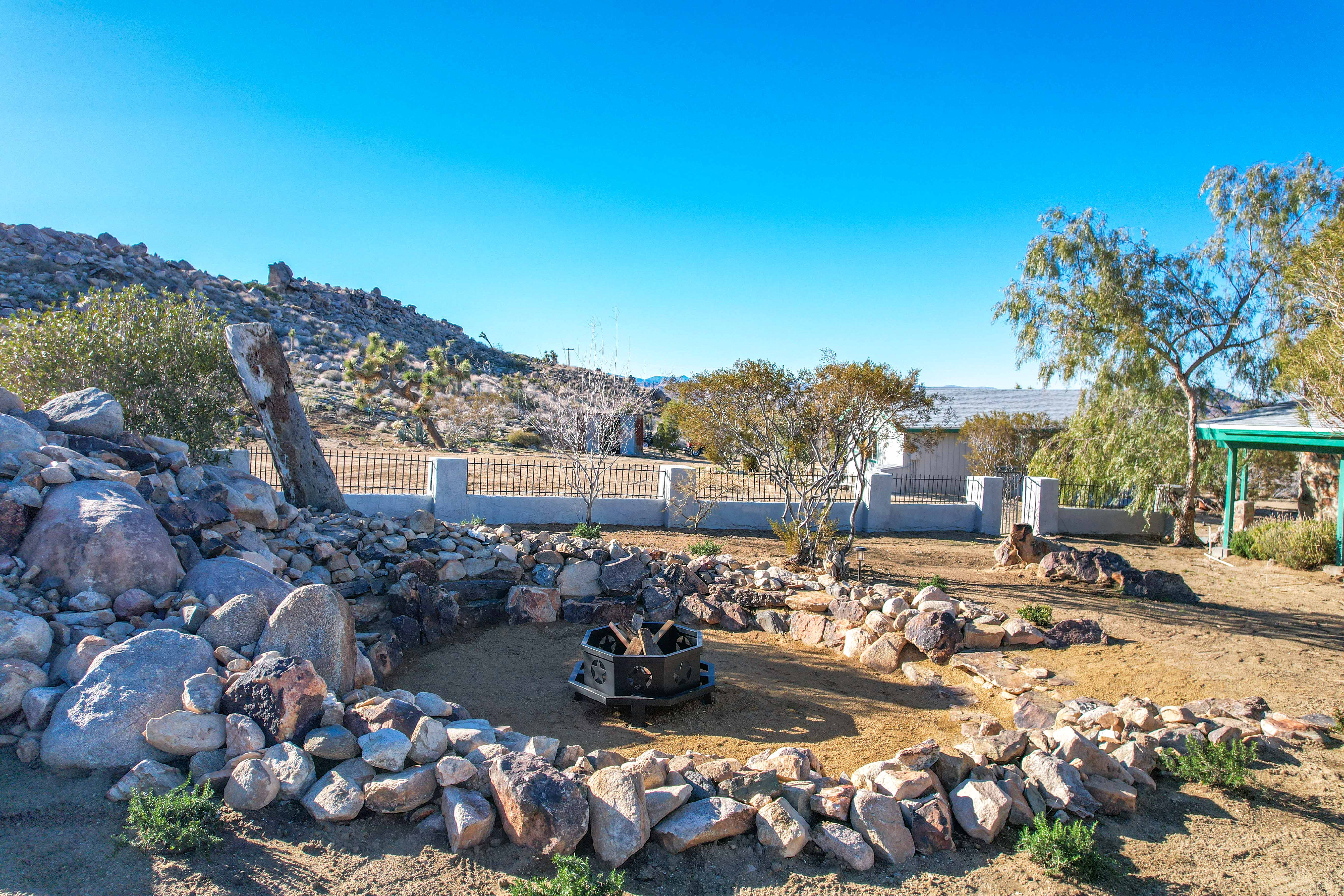 a Rustic Firepit Surrounded by Rocks at Onaga Oasis, with Desert Vegetation and Mountains in the Background Under a Clear Blue Sky.