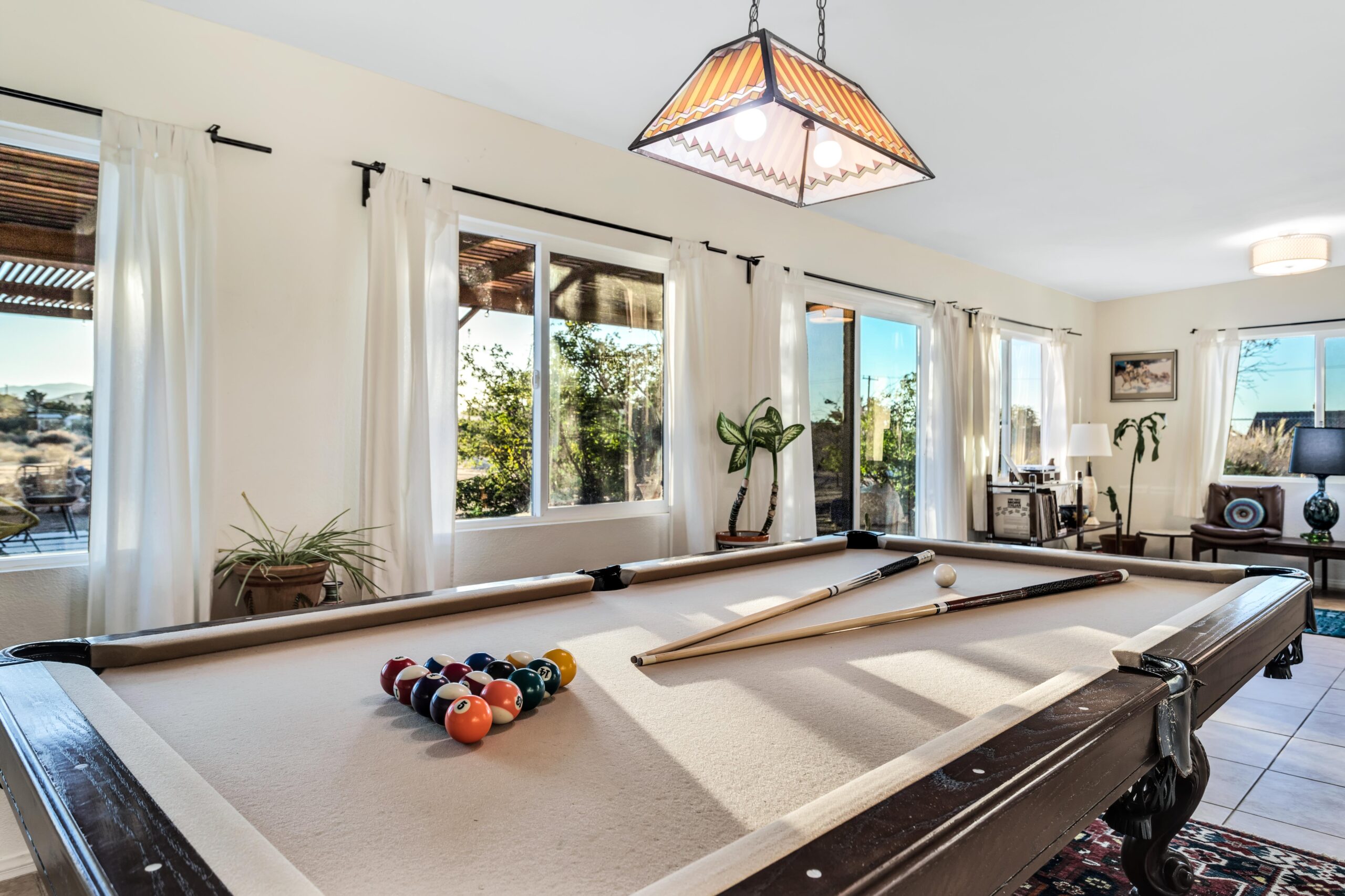 Sunlit Game Room with a Full-size Pool Table, Racked Billiard Balls, Cue Sticks, Large Windows with Desert Views, Record Player, and Board Games at Dez-art Retreat.