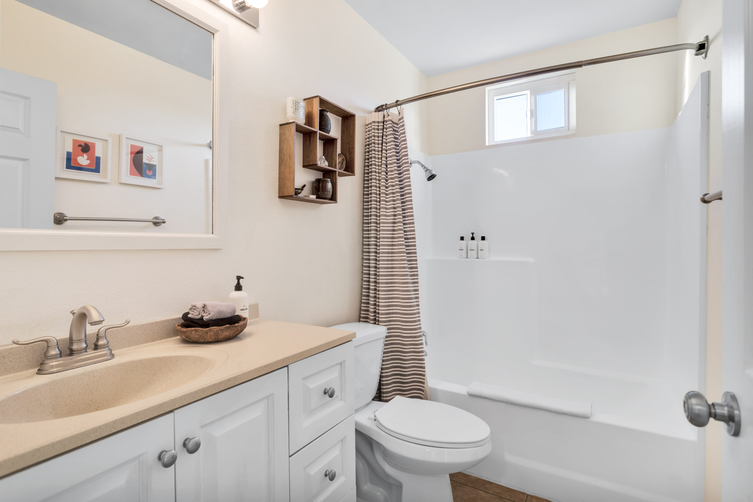 Bright Master Suite Bathroom with a White Tub and Shower Combo, Soft Neutral Walls, Open Shelving with Decor, Plush Towel, and Simple Modern Finishes at Dez-art Retreat.