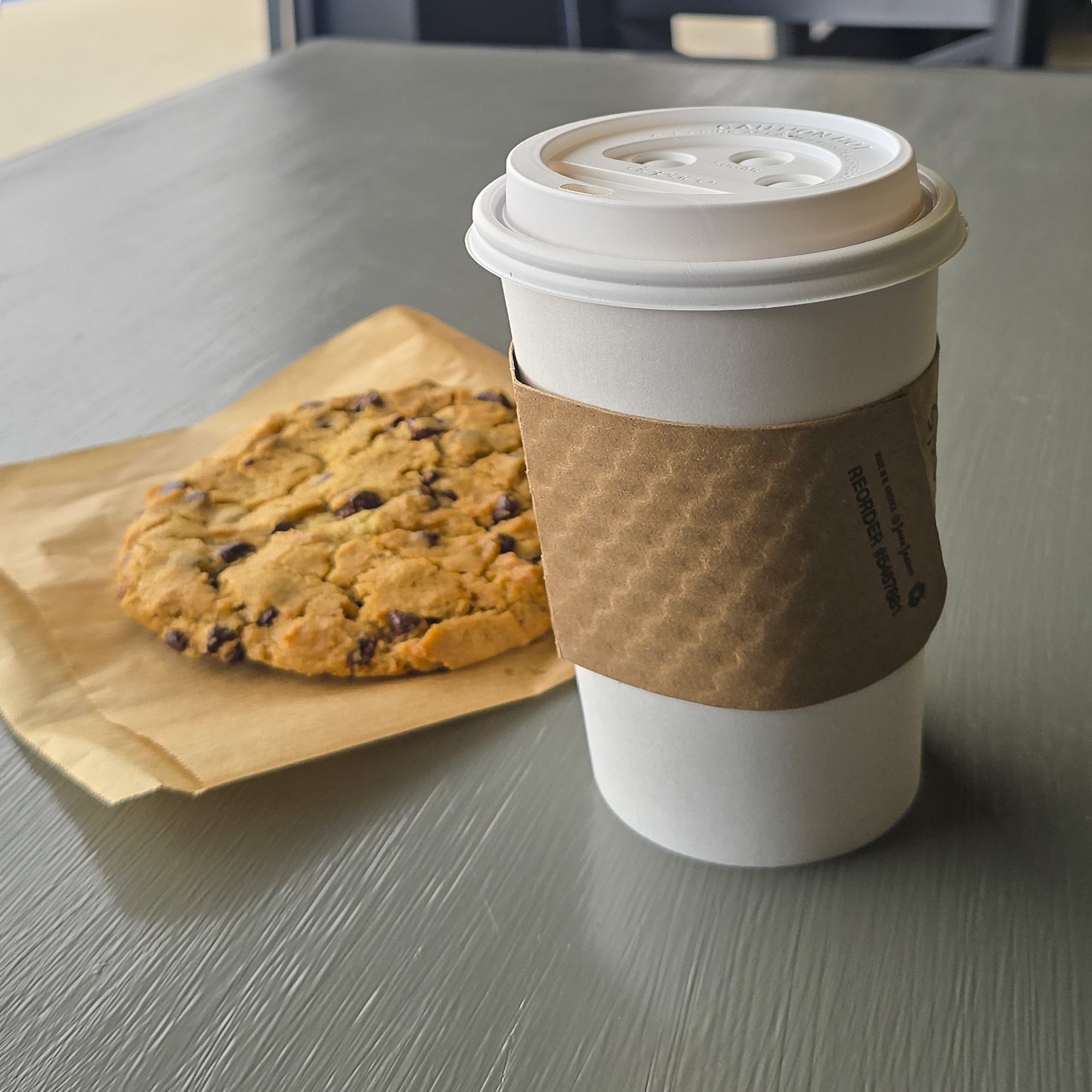 To-go Coffee Cup with Sleeve Beside a Chocolate Chip Cookie Resting on Parchment Paper on a Café Table at Bliss Cafe