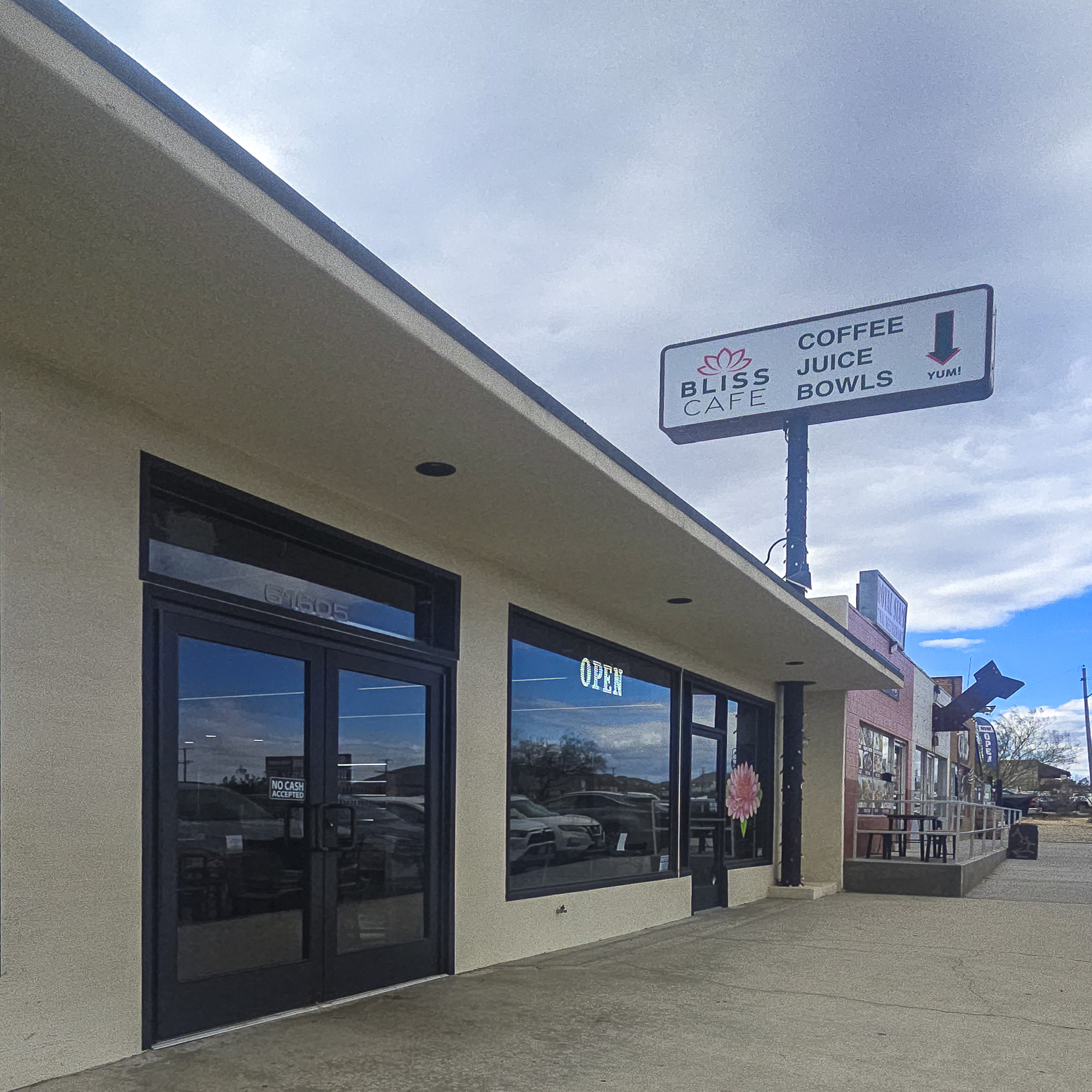 Daytime Exterior of Bliss Cafe in Joshua Tree, Showing the Storefront, Sidewalk Seating Area, and Sign Advertising Coffee, Juice, and Bowls.