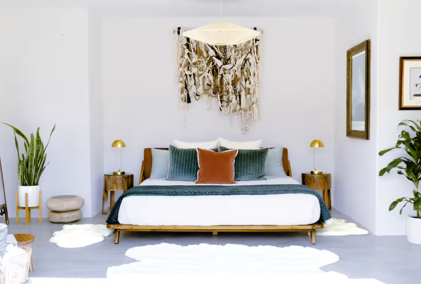 Primary Bedroom at Casa Coyote Featuring a Modern Wood Bed Frame with Layered Pillows, a Textured Wall Hanging Above the Bed, Matching Bedside Tables with Gold Lamps, Indoor Plants, and a Bright, Minimalist Desert-inspired Design.