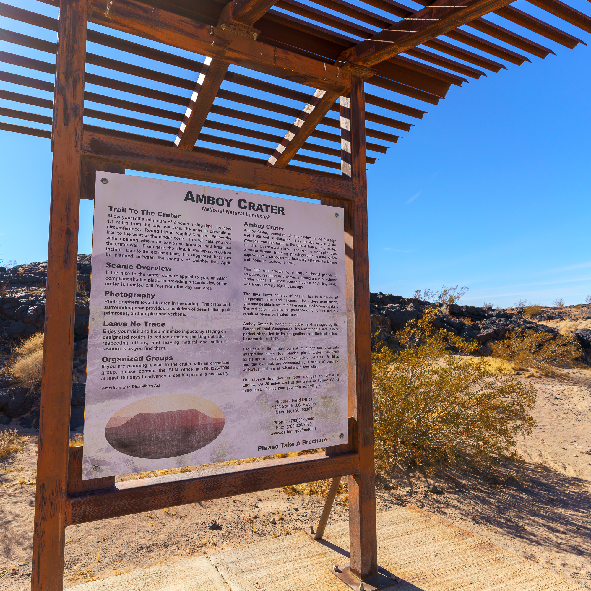 Trailhead Sign for Amboy Crater National Natural Landmark, Mounted Under a Wooden Shade Structure, with Desert Landscape, Volcanic Rocks, and a Clear Blue Sky in the Background.