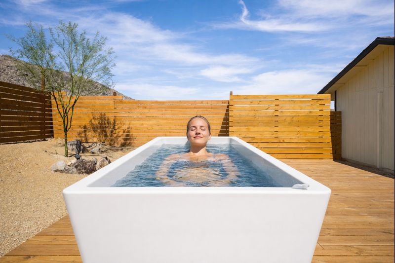 Ai-generated Image of a White Cold Plunge Tub Positioned Lengthwise on a Wooden Deck in the Desert, with a Woman Relaxing Inside the Tub, Eyes Closed, Framed by a Wooden Privacy Fence and a Bright Blue Sky.